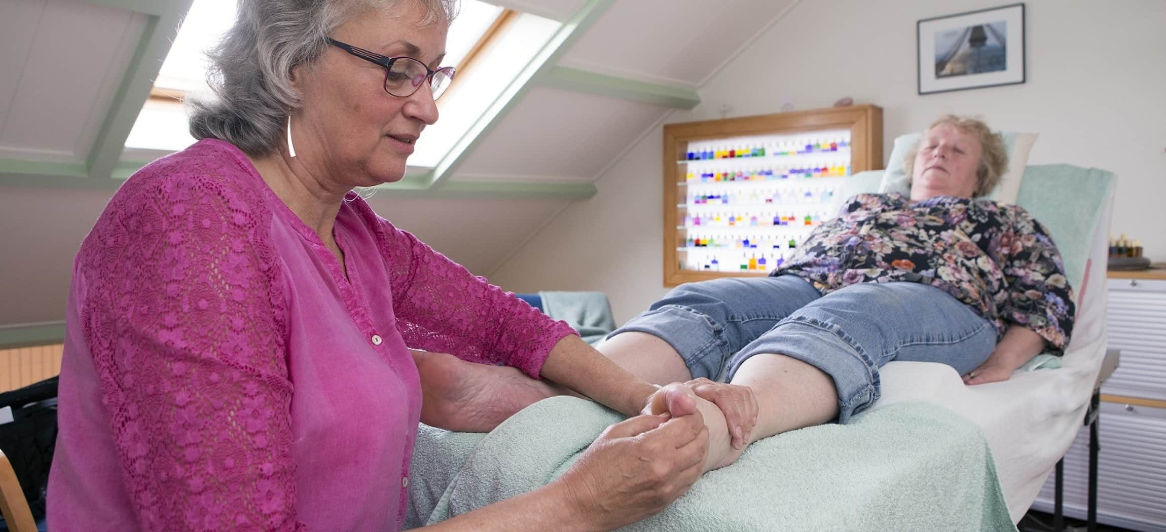 Vrouw met roze blouse masseert de voet van een vrouw die op een massagetafel ligt