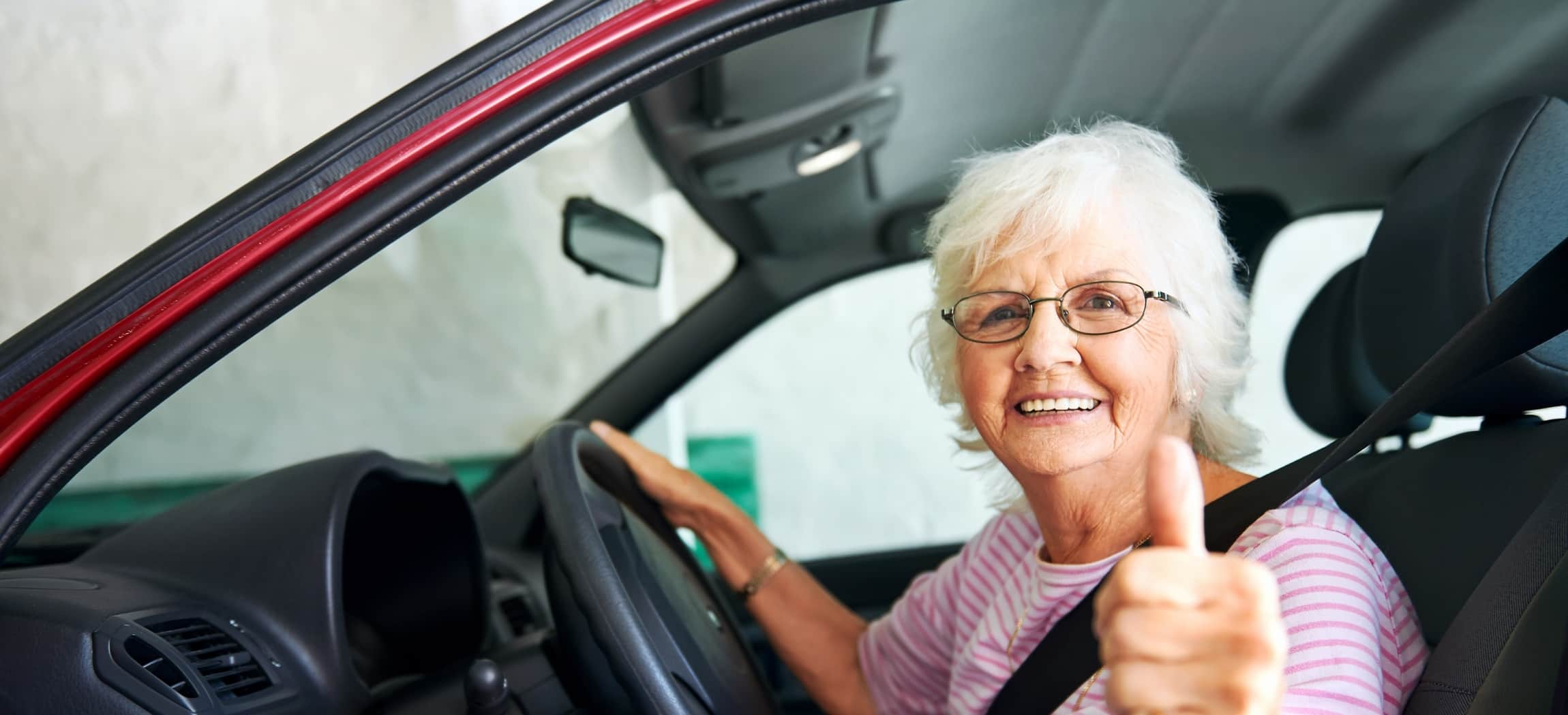 Vrouw grijs haar met bril zit achter het stuur in een auto met duim omhoog