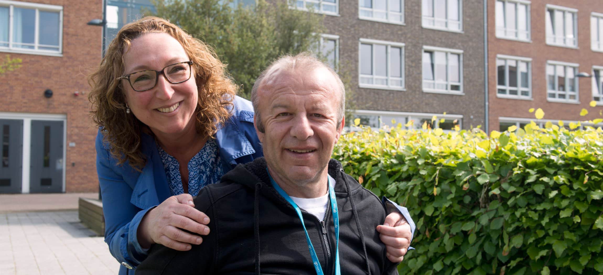 Jonge vrouw kijkt over de schouder van een man die in een rolstoel zit, groene heg en huizen op achtergrond