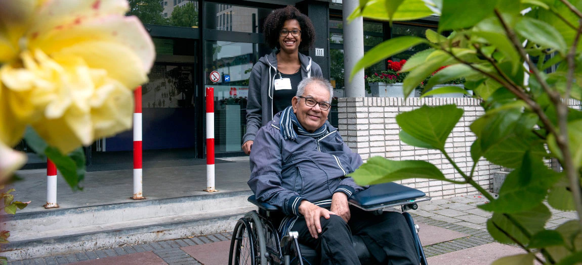 Man in rolstoel voor de deur van een verpleeghuis met een verpleegkundige