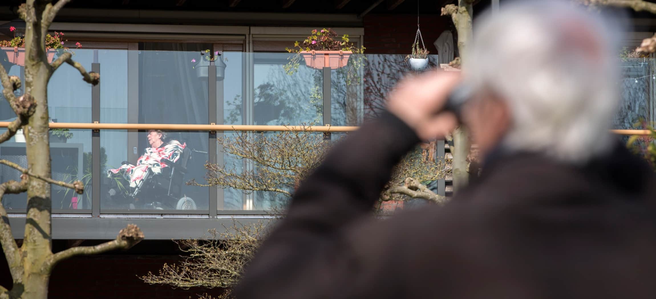 Oudere heer kijkt met verrekijker naar zijn vrouw op het balkon van het verpleeghuis