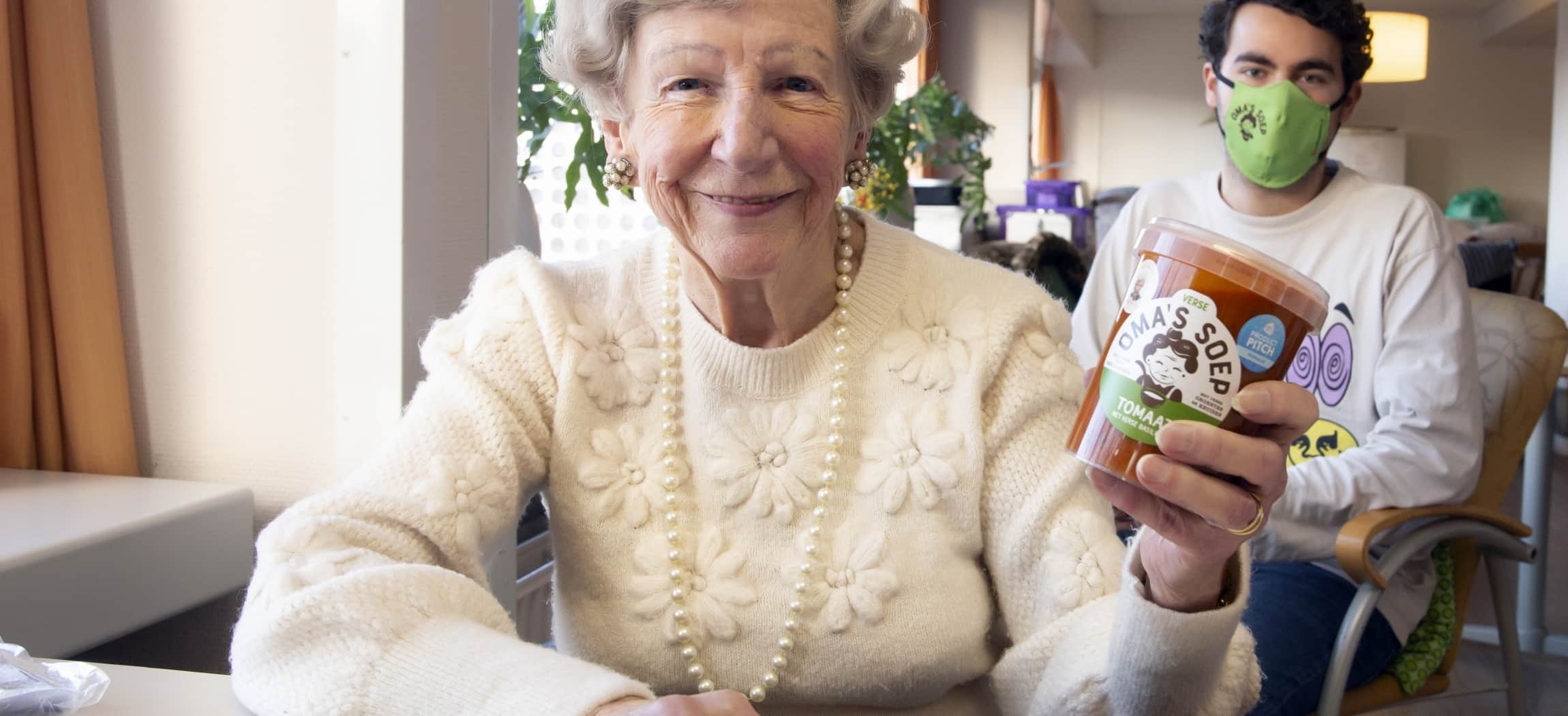 Oudere dame toont pot 'oma's soep' en jonge man zit achter haar