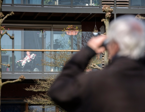Oudere heer kijkt met verrekijker naar zijn vrouw op het balkon van het verpleeghuis