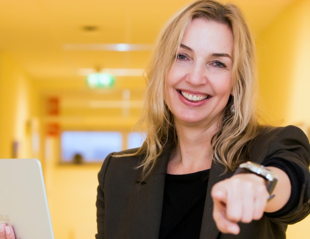 Vrouw met blonde haren in zwart colbertjasje houdt Chromebook vast en wijst naar de camera in gele gang